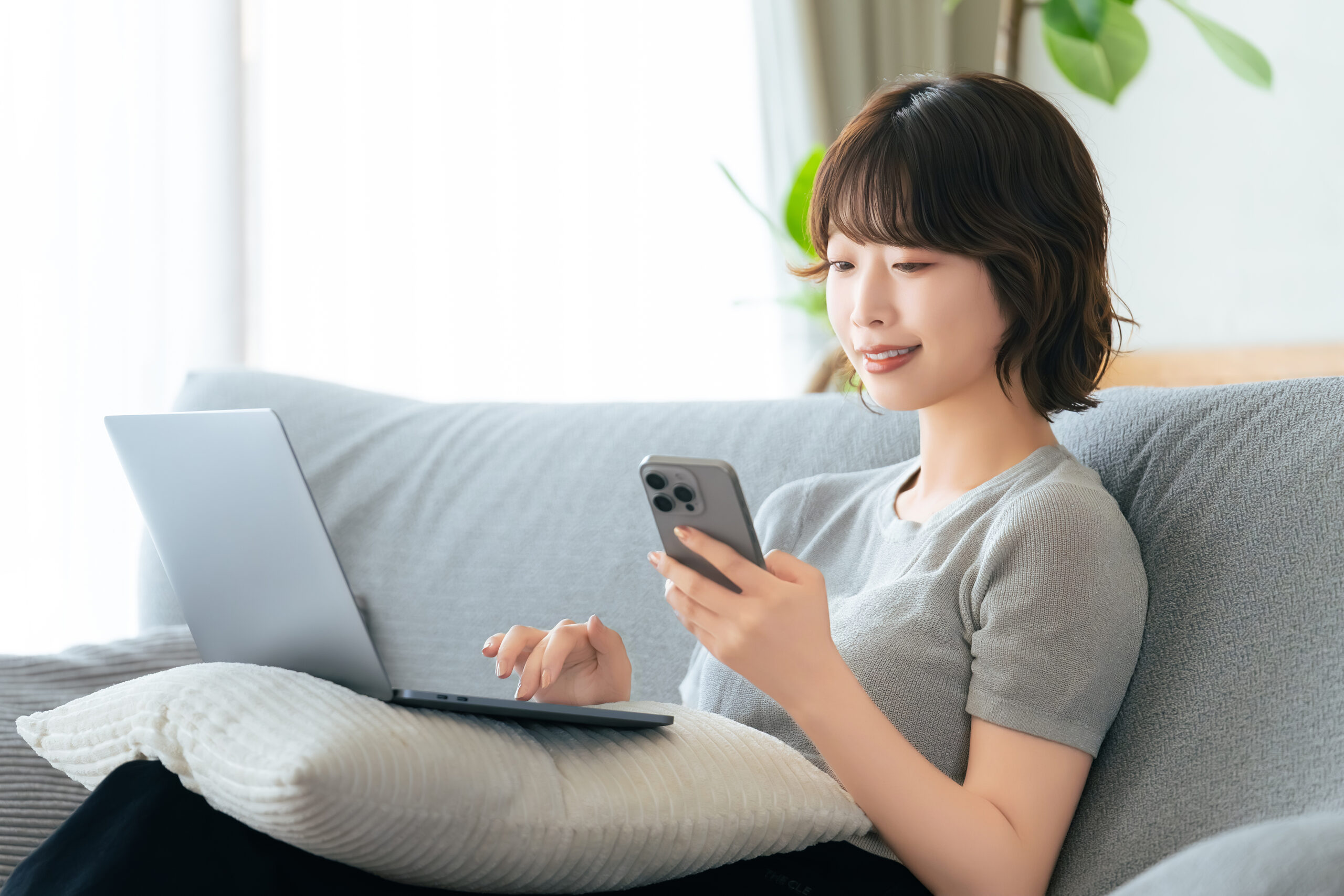 ソファに座ってスマホとパソコンを使う女性